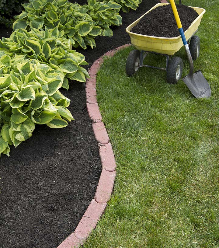 A garden scene showing a neatly edged flower bed with green hostas and dark mulch. A yellow wheelbarrow filled with mulch and a shovel are positioned on a well-maintained lawn beside the bed's brick border.