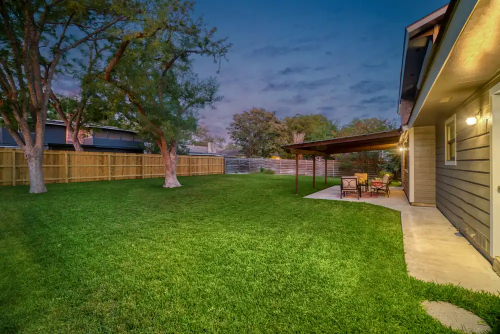 A spacious backyard at dusk features green grass, a large tree, a wooden fence, and a covered patio area with outdoor furniture next to a house. The sky is partly cloudy with soft evening light.