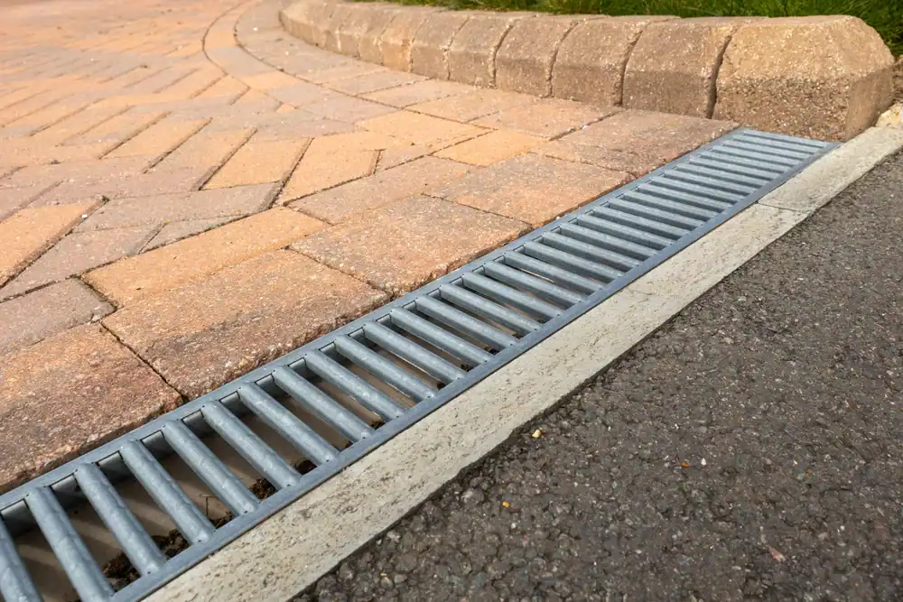 Metal drainage grate installed between an asphalt road and a brick-paved sidewalk, with a raised curb and some grass visible at the top edge of the image.