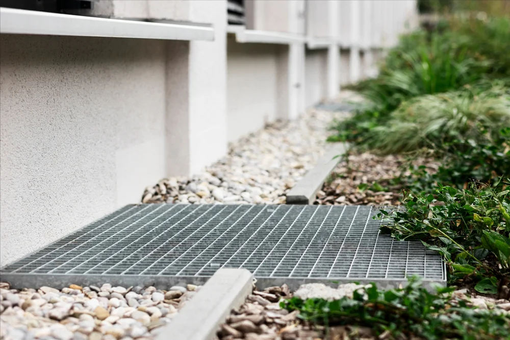 A close-up view of a metal grate on the ground near a building, surrounded by small rocks, concrete edging, and green plants along the side.