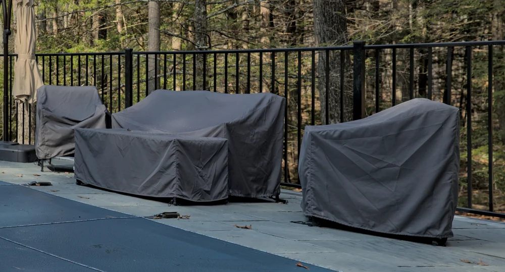 Three pieces of outdoor furniture covered with gray protective covers are lined up on a patio next to a black metal fence, with trees visible in the background.