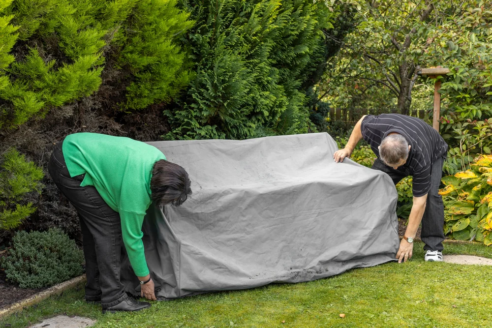 Two people are covering an outdoor bench with a large gray protective cover in a garden surrounded by green shrubs and plants.