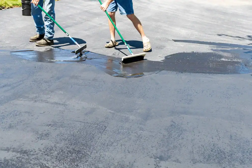 Two people use long-handled tools to spread sealant on a driveway, resurfacing and protecting it. One wears jeans and the other denim shorts. Only their legs and tools are visible.