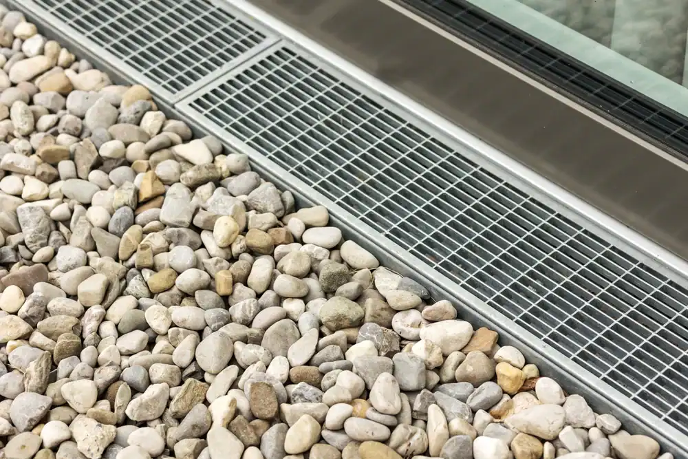 Metal drainage grate next to a building’s glass wall, bordered by a section of small, smooth, light-colored stones.