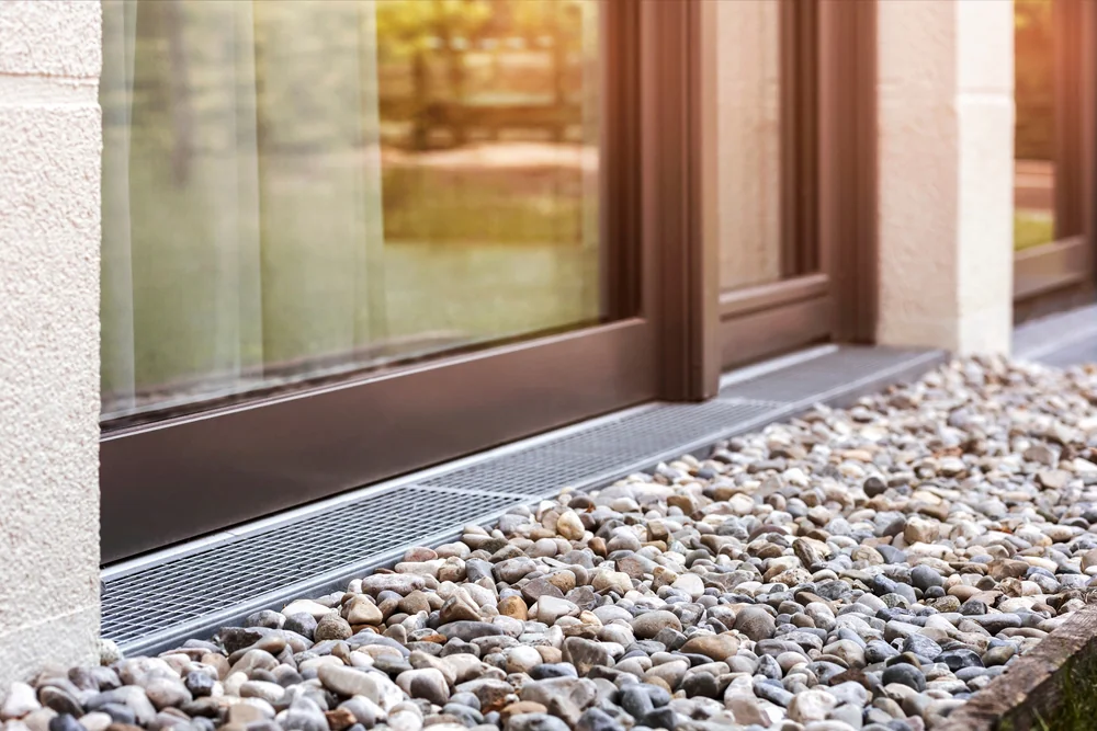 Close-up of a modern glass door with a dark frame, next to a building wall. In front of the door, there is a metal drainage grate surrounded by small decorative pebbles. Sunlight reflects off the glass.