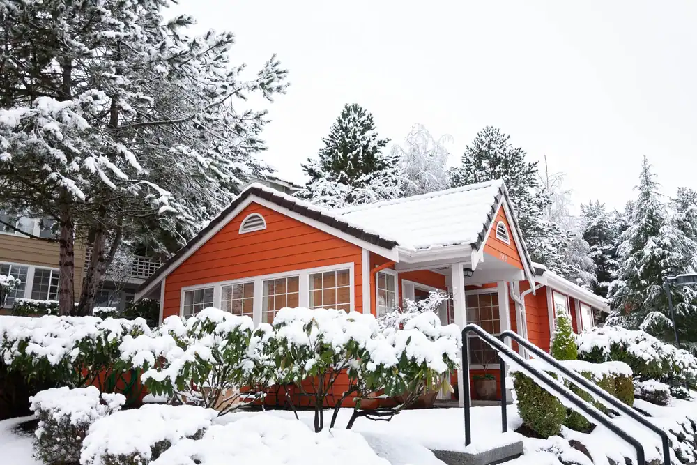 A bright orange house with white trim is surrounded by snow-covered trees and bushes. Snow blankets the roof, yard, and steps leading to the front door, creating a wintery scene.
