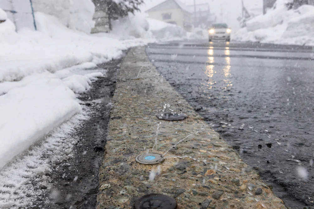 Low angle view of a wet street in winter, with water spray nozzles embedded in the curb, snow piled on both sides, and a car with headlights on approaching in the background during snowfall.