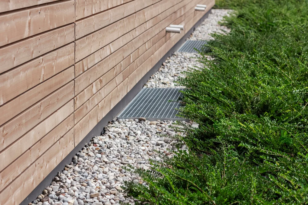 Metal grates and white pebbles border the base of a wooden exterior wall, with green shrubs growing alongside, creating a neat separation between the building and the landscaping.