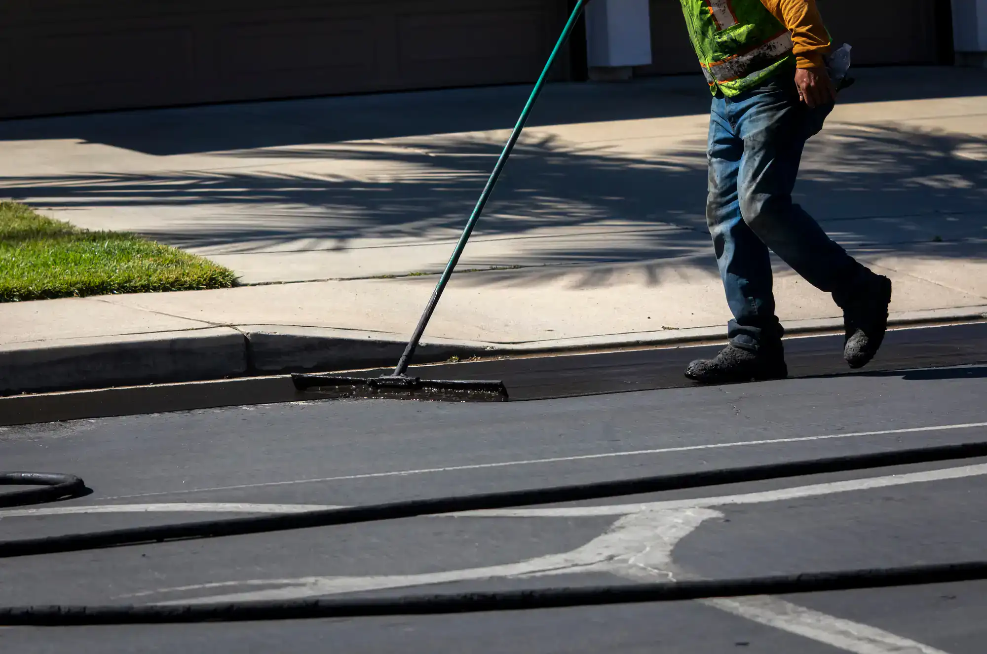 A worker in a safety vest and boots uses a long-handled tool to spread asphalt or sealant on a suburban street near a curb on a sunny day.