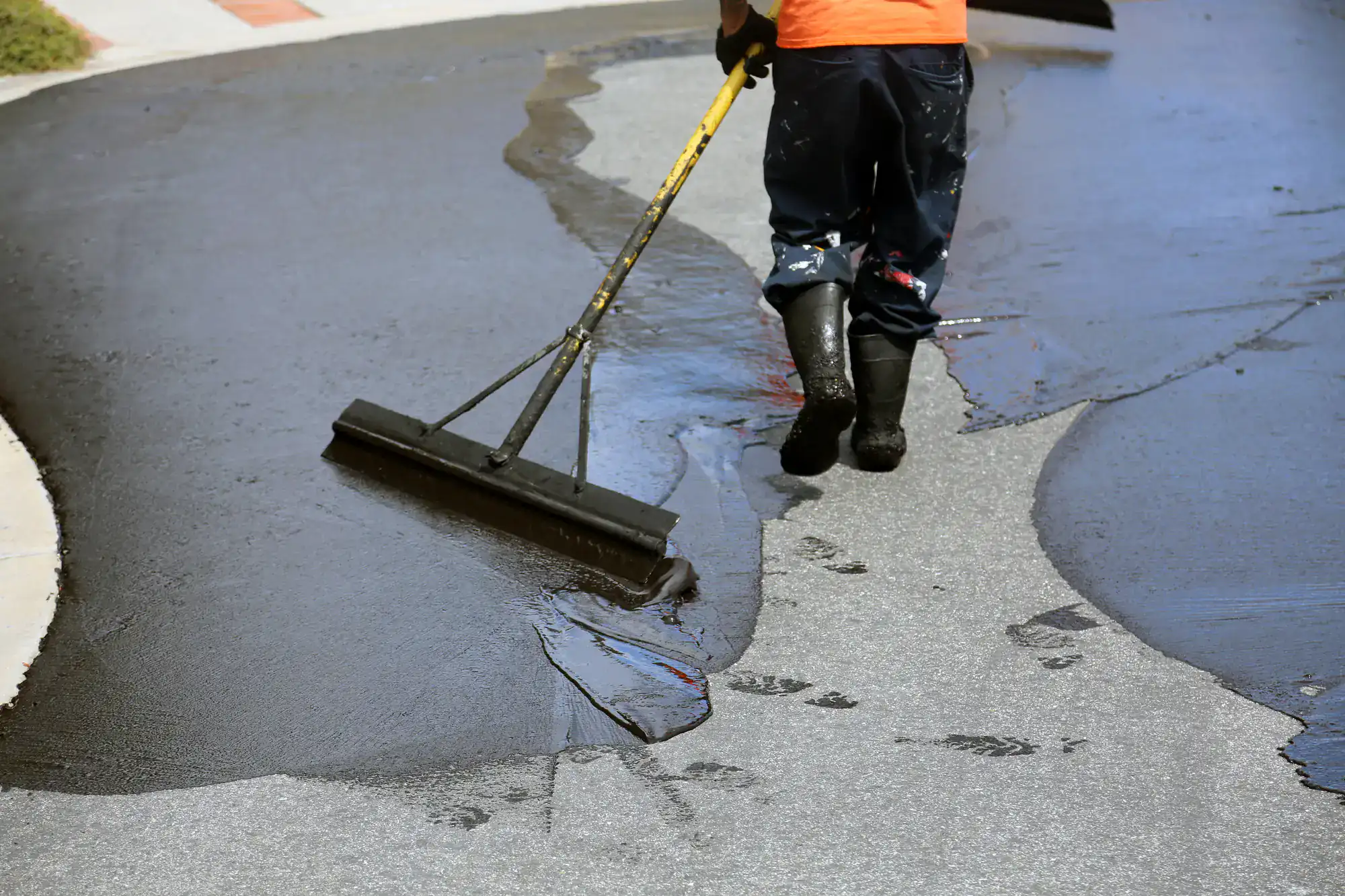 A person wearing boots spreads black sealant on a driveway with a large squeegee, leaving footprints on the wet surface as they work.