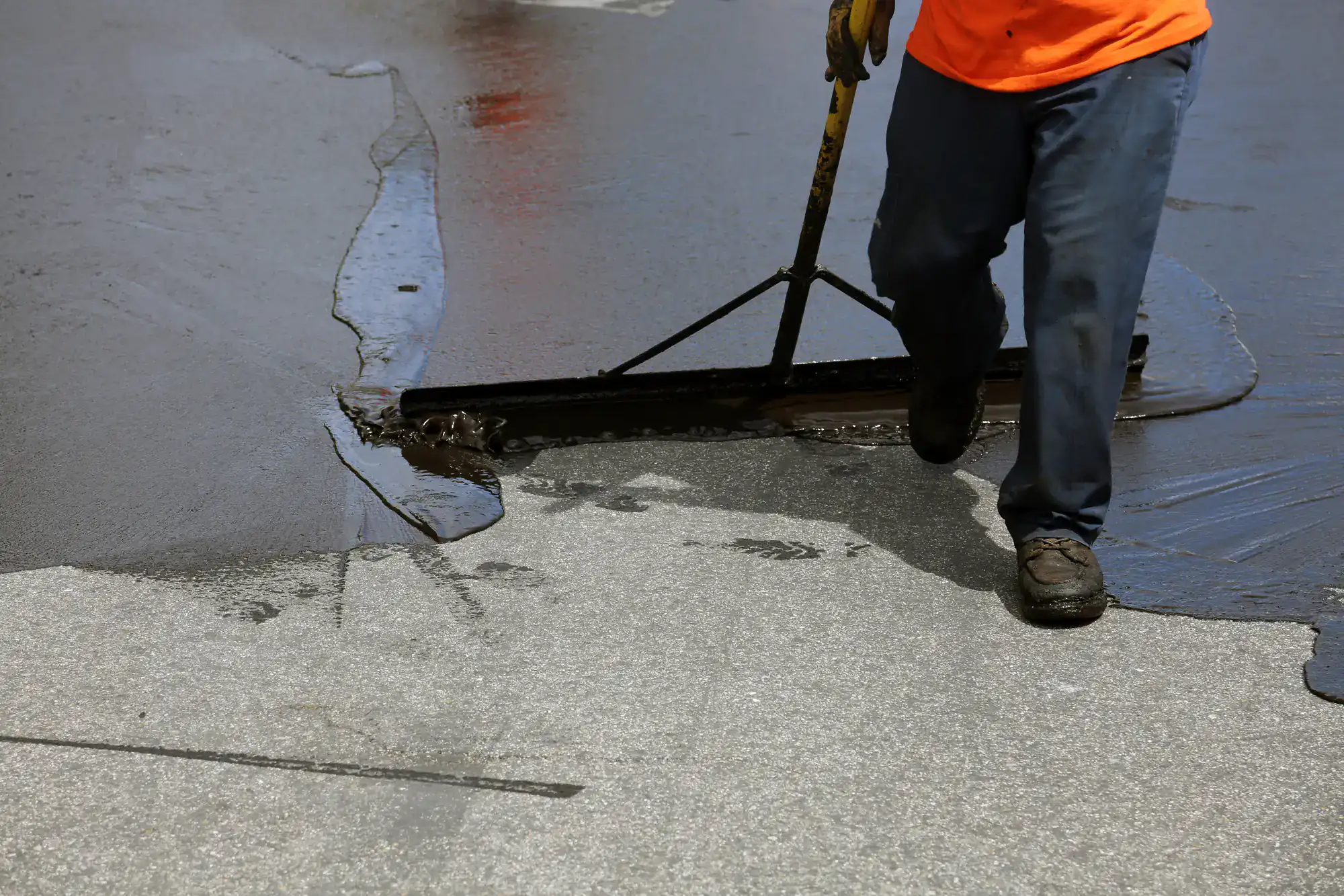 A worker in an orange shirt uses a large rake to spread black sealant over a gray asphalt surface, resurfacing the pavement. Only the worker’s lower body is visible.