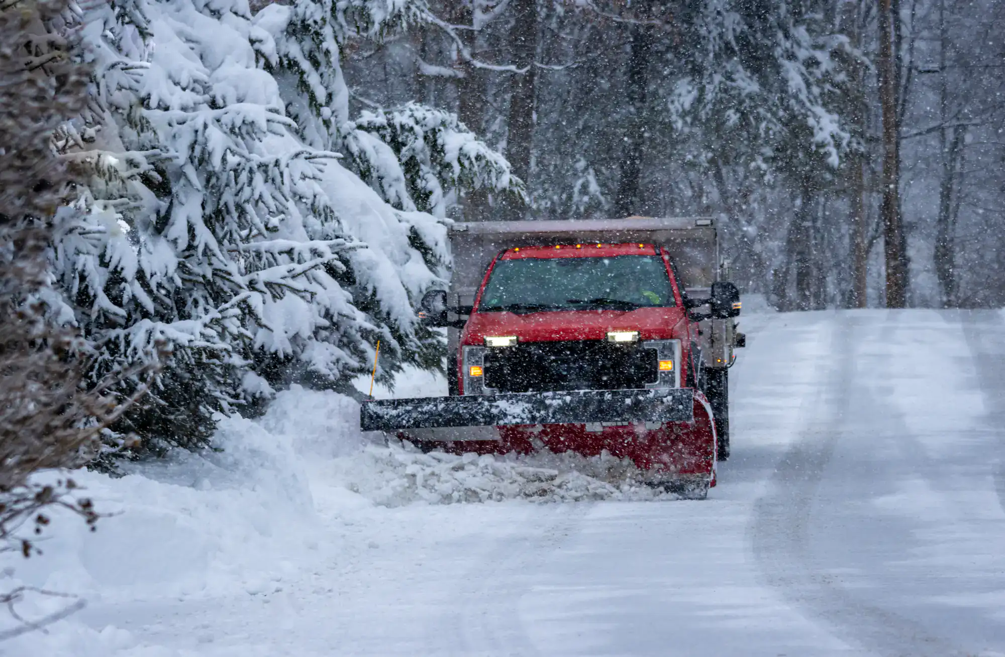 A red snowplow truck clears snow from a tree-lined road during heavy snowfall, with snow-covered trees and a wintry scene in the background.