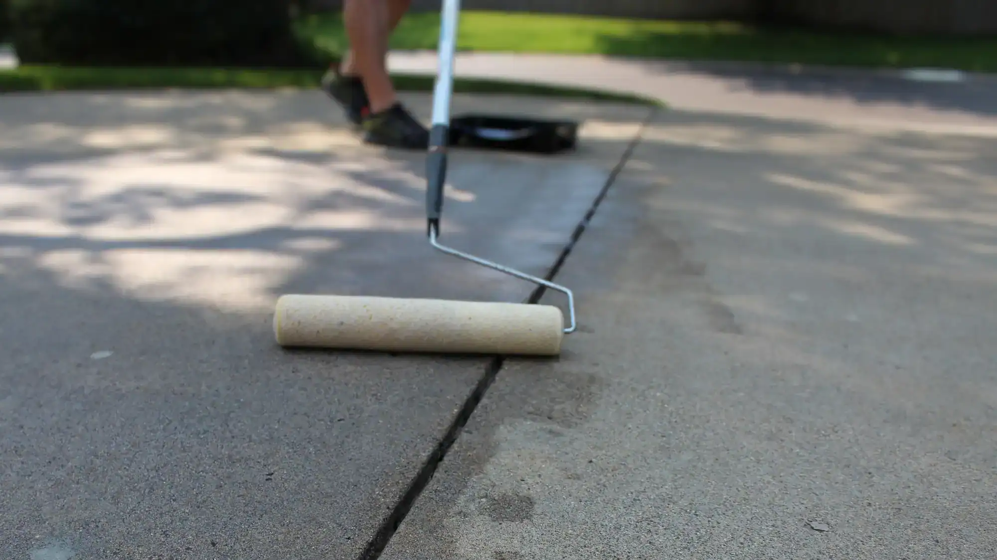 A close-up of a paint roller being used to apply sealant to a concrete driveway. A person’s legs are visible in the background, along with a paint tray and green grass.