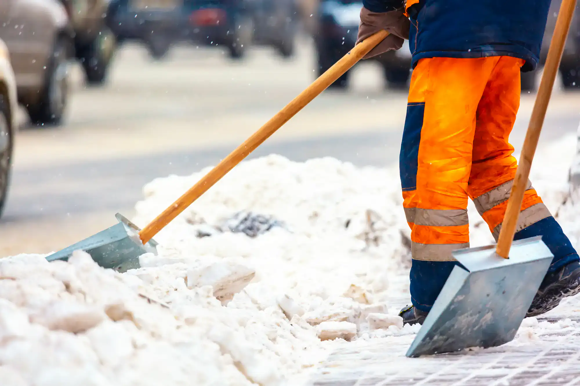 Two people in bright orange work pants use shovels to clear snow from a sidewalk. Snow piles are visible, and a road with blurred cars is in the background.