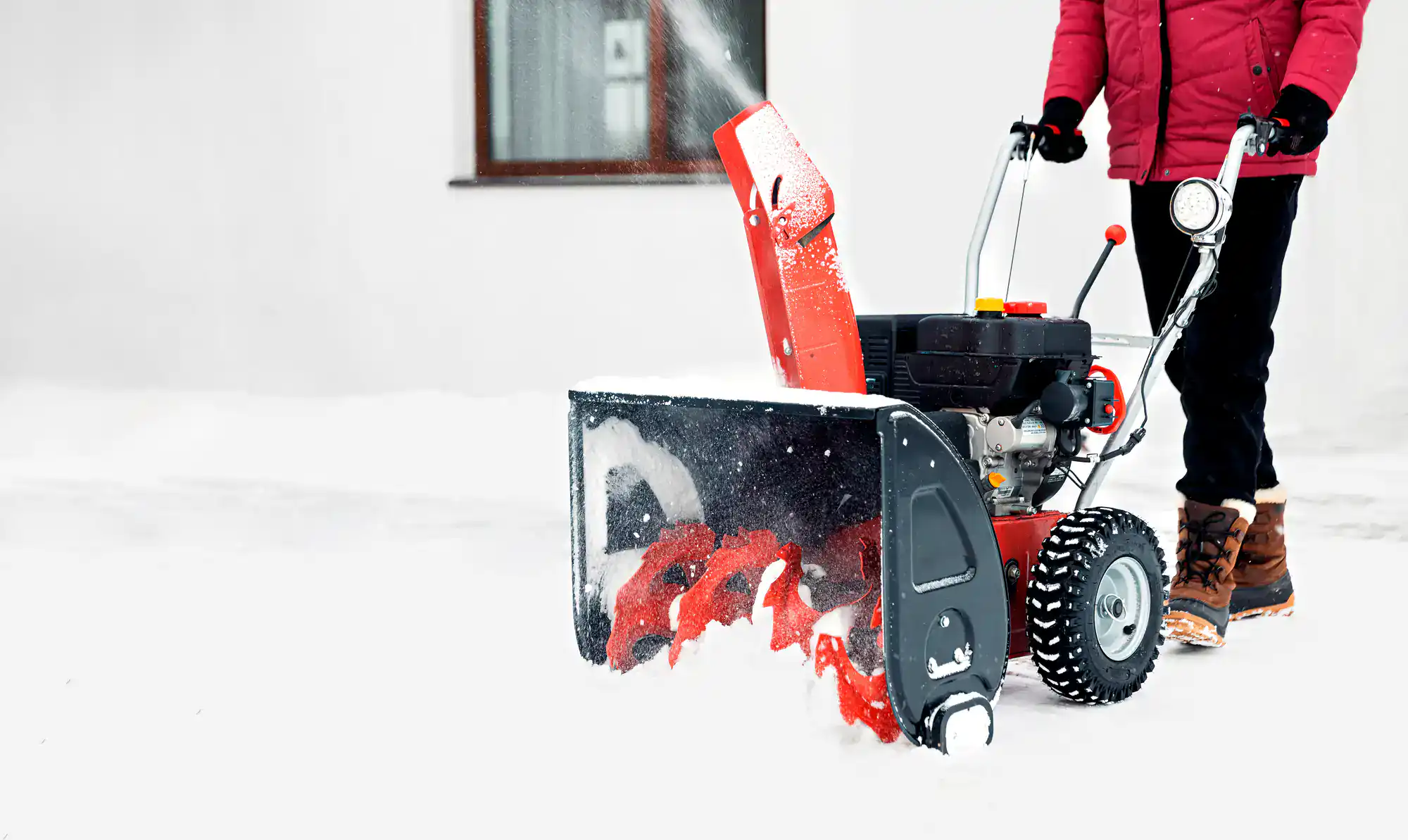 A person in a red jacket and winter boots operates a red snow blower, clearing fresh snow from a driveway in front of a white building with a window.