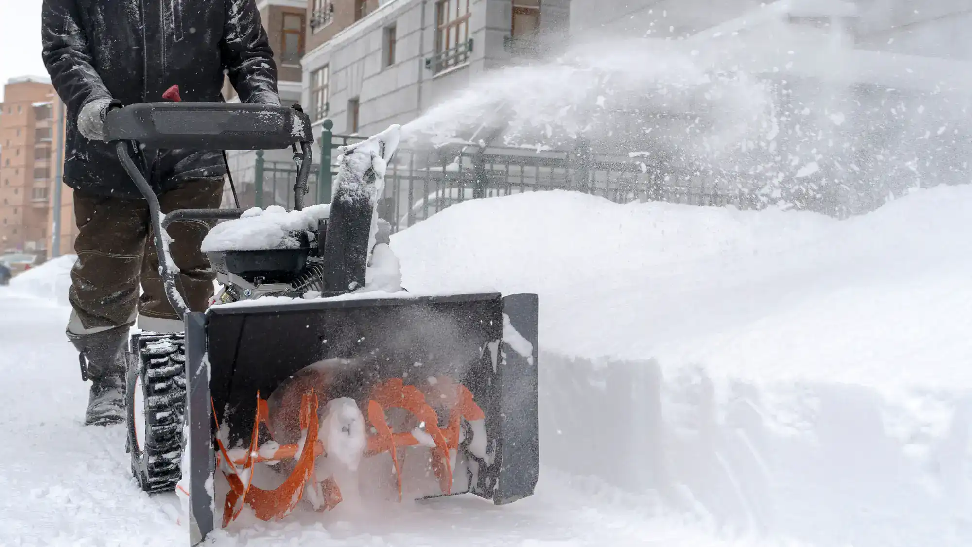 A person uses a snow blower to clear a thick layer of snow from a sidewalk in front of buildings on a winter day. Snow is blowing out of the machine in an arc.