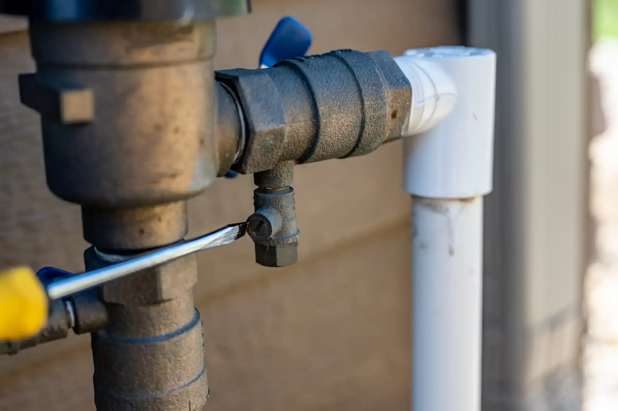 A close-up of a screwdriver turning a screw on a metal outdoor water pipe next to a white PVC pipe, against a brown wall background.