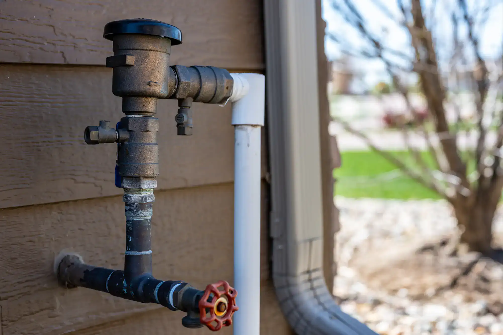 A backflow preventer with a red valve is attached to the exterior wall of a house, next to a downspout and surrounded by landscaping with rocks and a tree in the background.