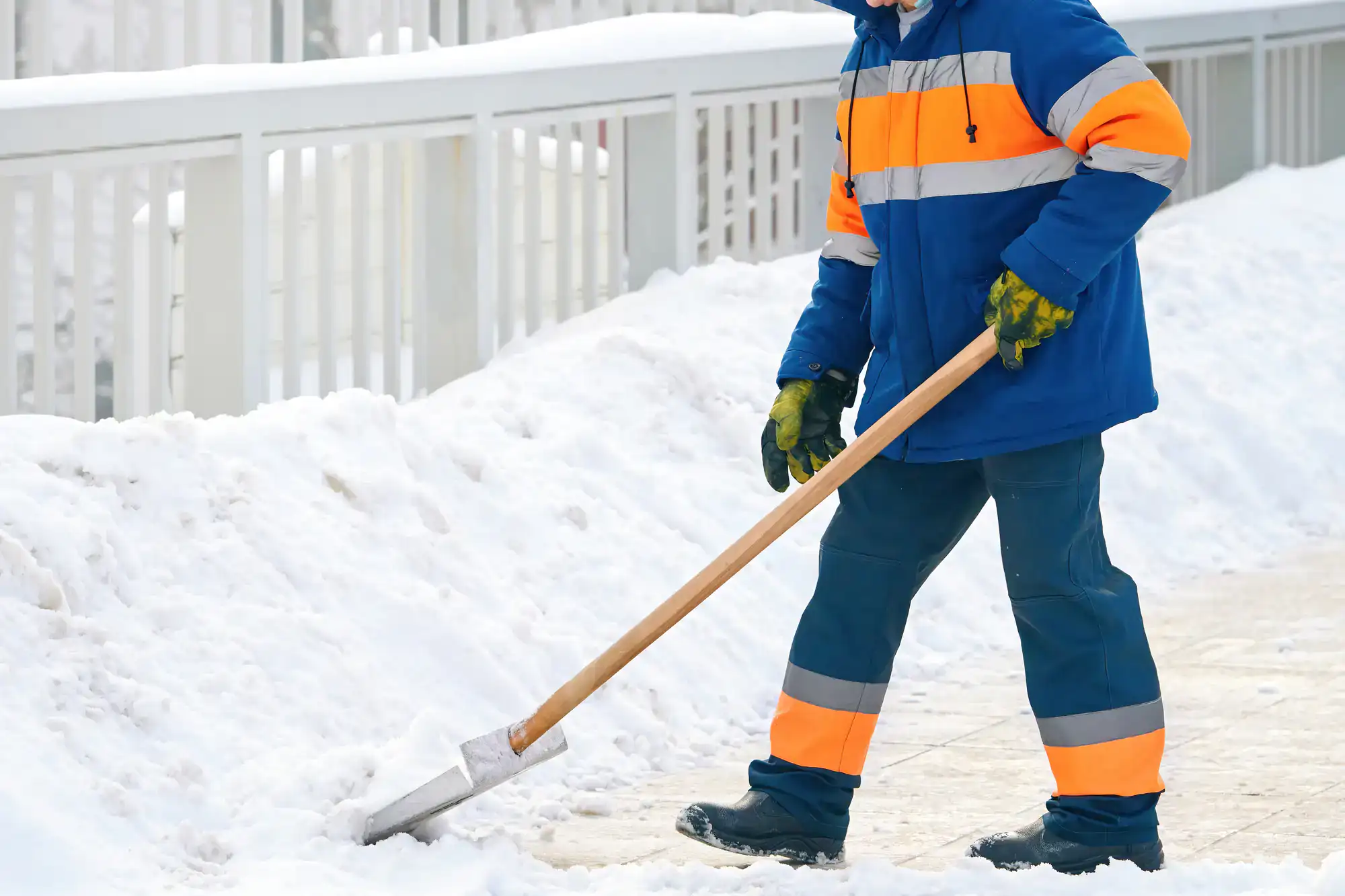A person wearing a blue and orange reflective winter uniform and green gloves is shoveling snow from a sidewalk next to a white fence.