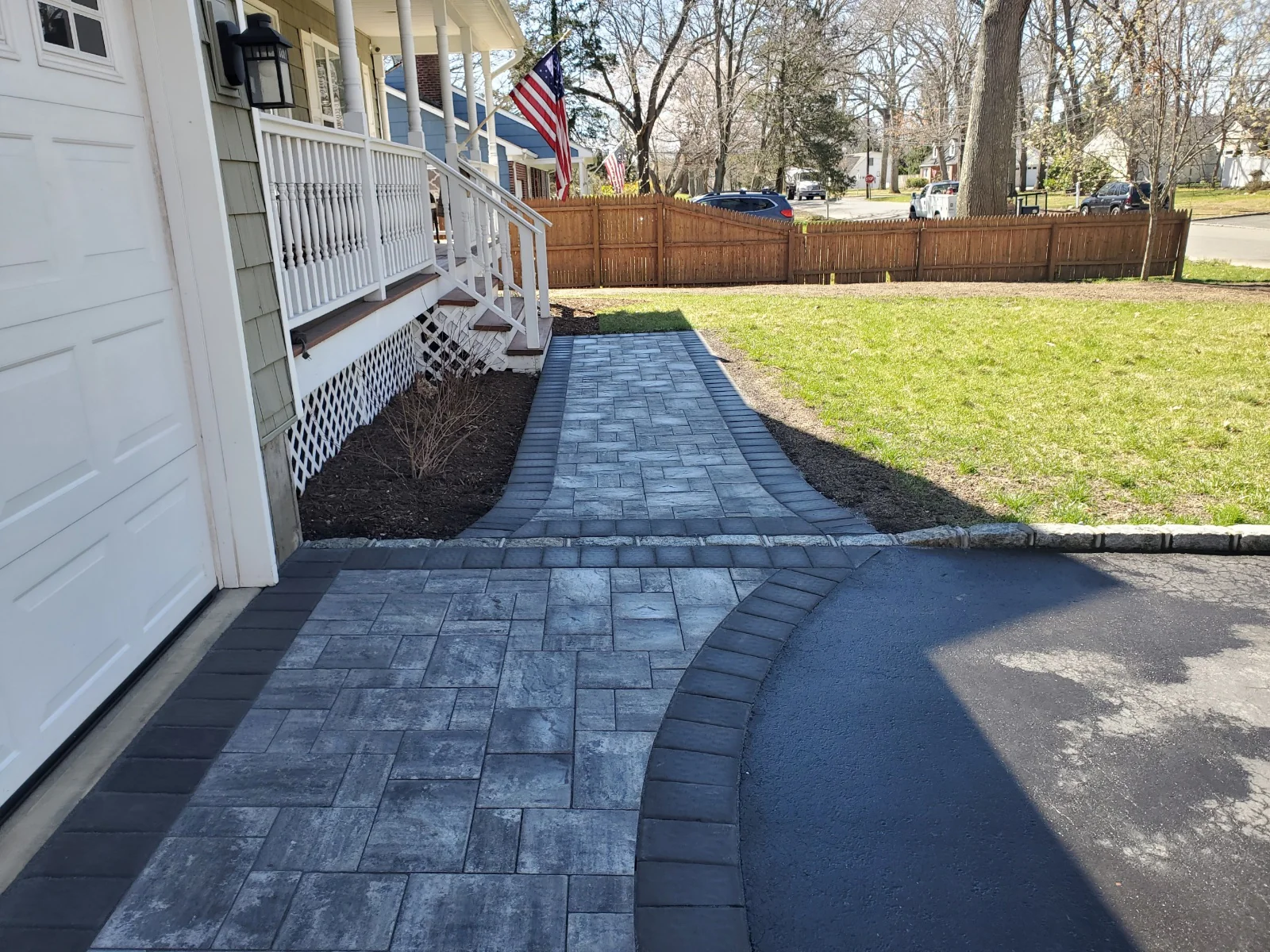 A paved stone walkway leads from a driveway to a front porch with white railings. The path curves through a yard with green grass, bordered by a wooden fence and trees in a suburban neighborhood.