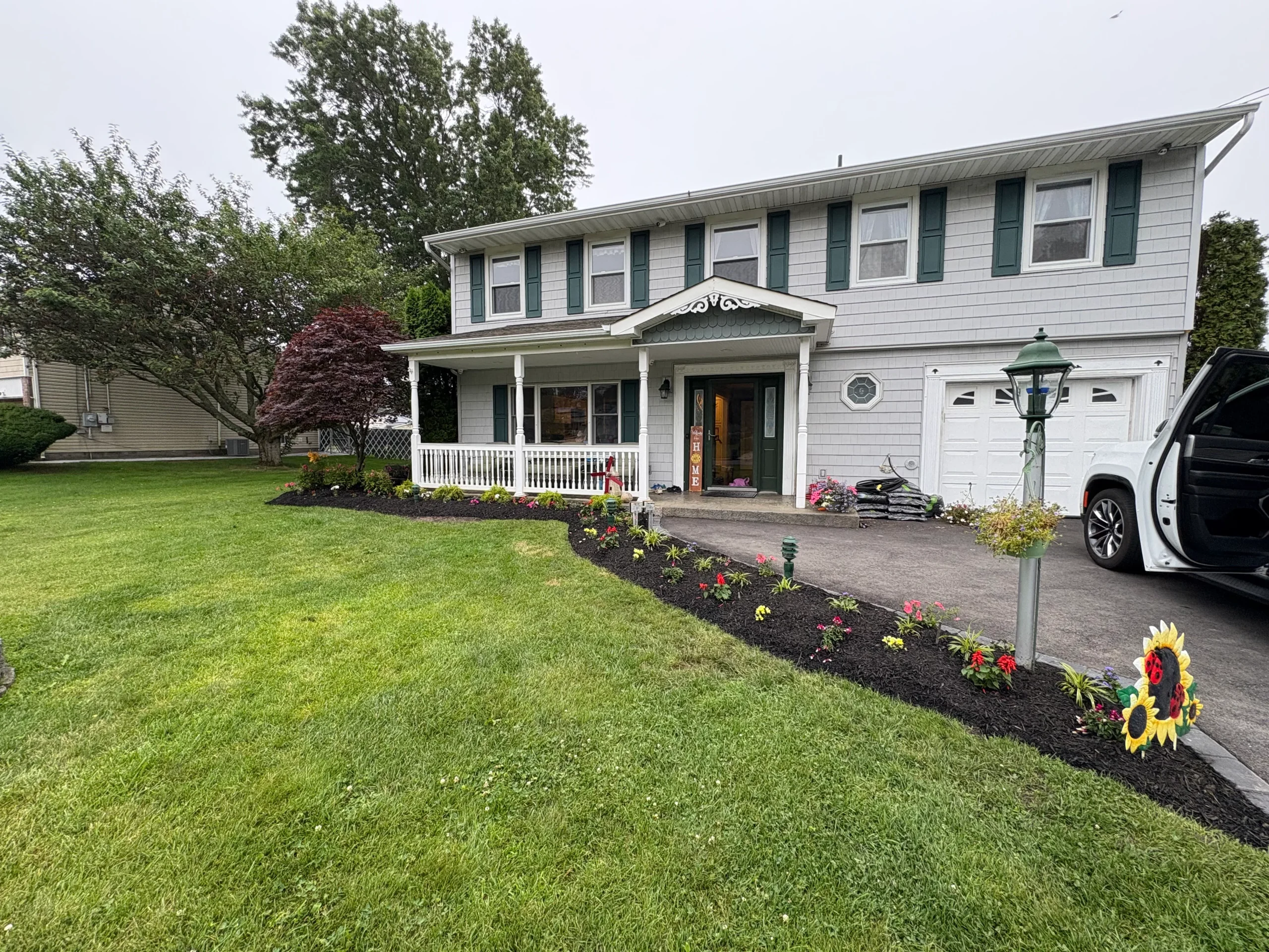 A two-story house with gray siding, white trim, a front porch, and green shutters, surrounded by a well-kept lawn and a flower bed. A car with an open trunk is parked in the driveway.