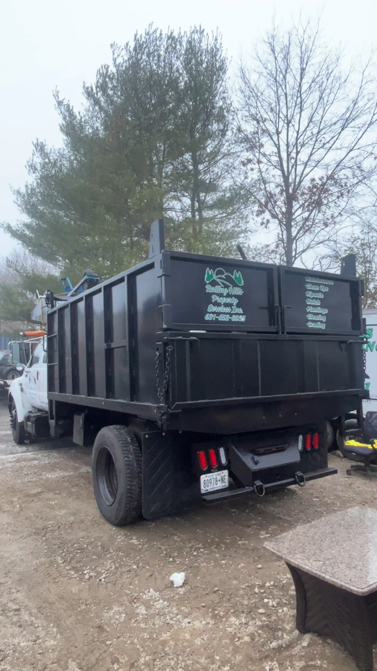 A large black dump truck with company info on the back is parked outdoors near leafy trees on a cloudy day.