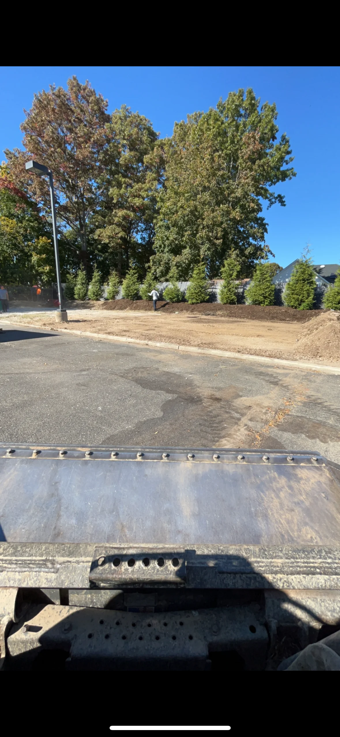 View from construction vehicle of a newly paved area by trees, shrubs, and blue sky at construction site.