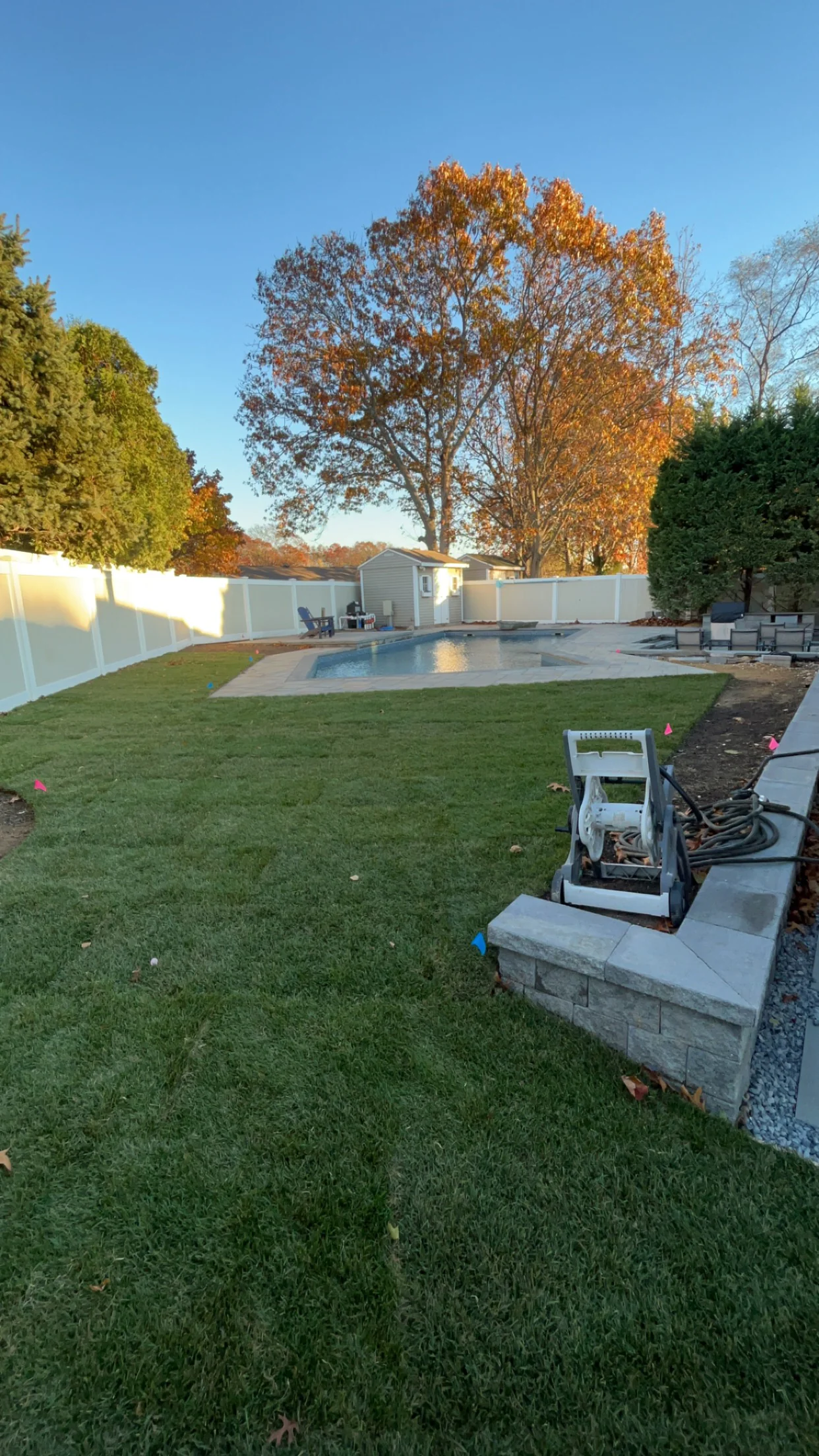 A backyard features fresh sod, a pool, stone patio, and landscaping equipment beside a retaining wall.