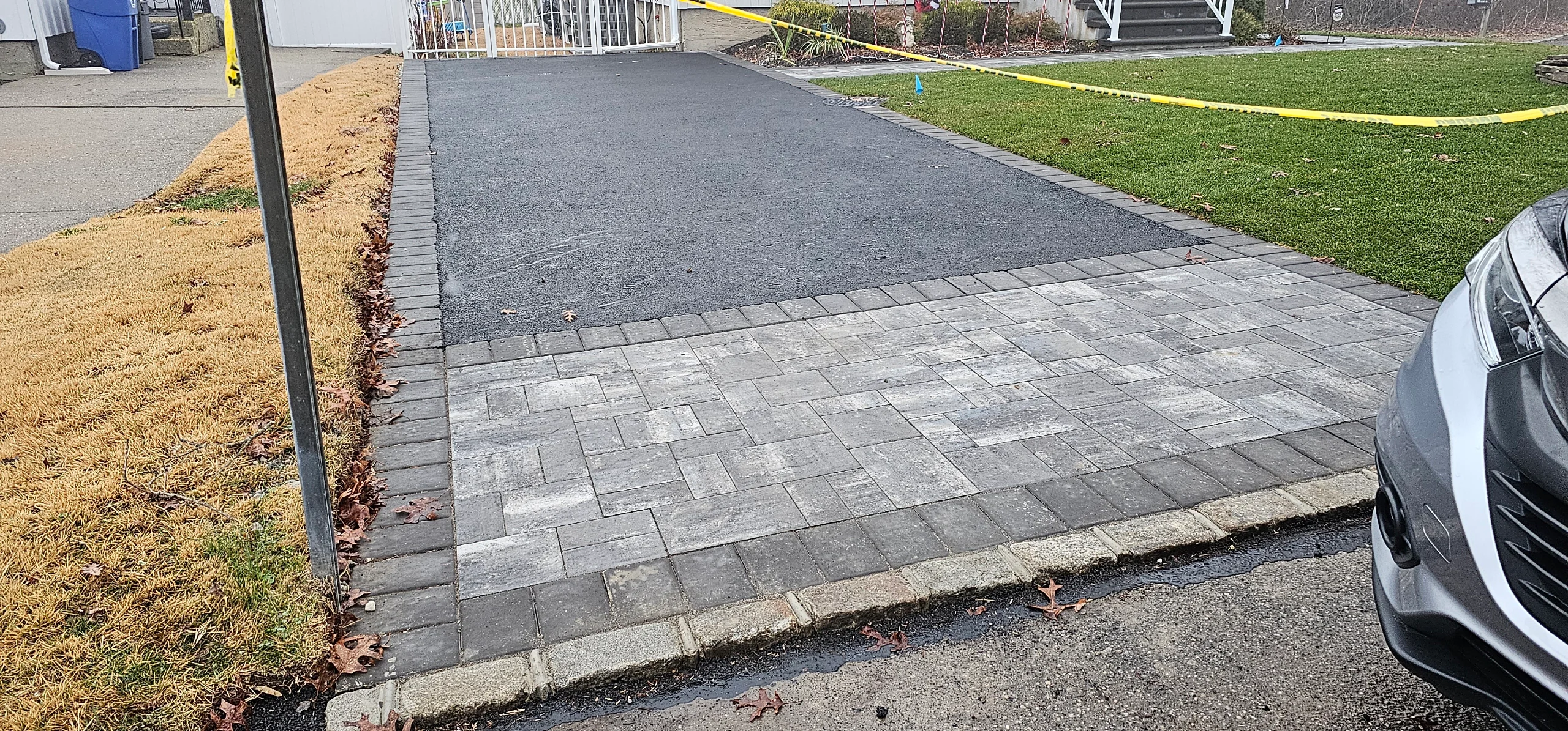 A freshly paved driveway with stone pavers at the entrance is bordered by grass, curb, and yellow caution tape.