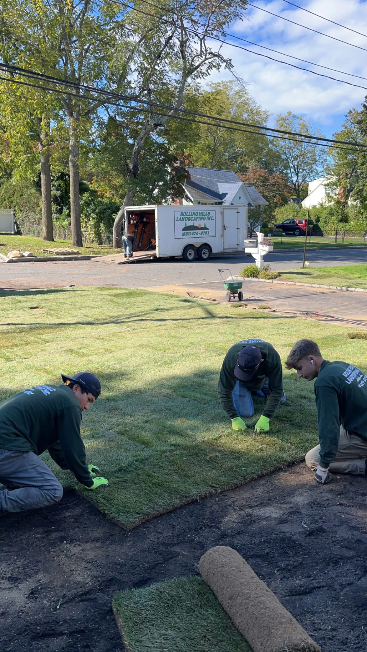 Three workers install sod on a lawn using landscaping equipment, with a trailer visible in the background.