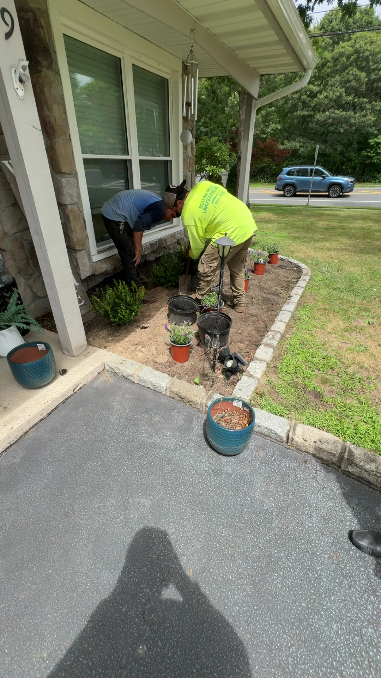 Two people are gardening by a house, planting shrubs and flowers in a bordered soil bed near the driveway.