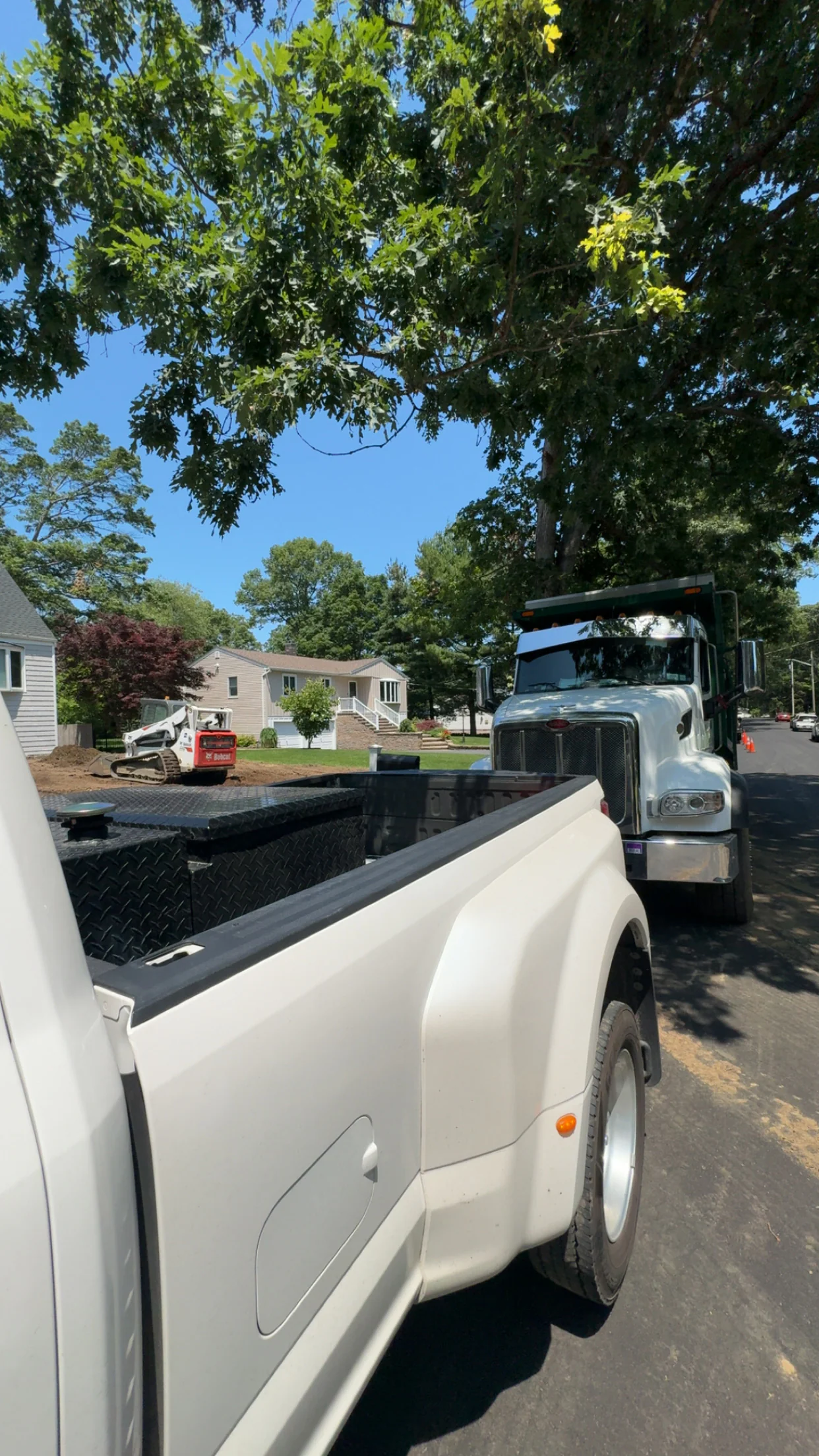 A white pickup truck and a large commercial vehicle are parked on a quiet suburban street with houses and trees.